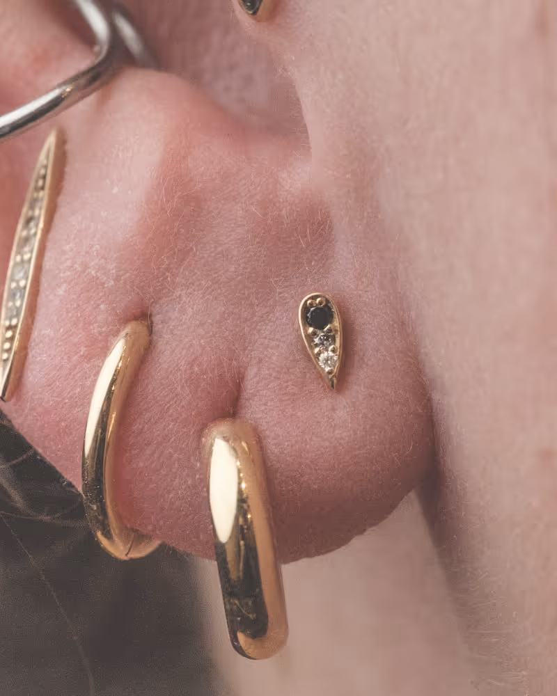 Close‑up of an ear wearing multiple gold earrings: two stacked hoops, a slender bar with pavé diamonds and a teardrop‑shaped stud with a dark centre stone and clear accents – naughtycomrade.ca.