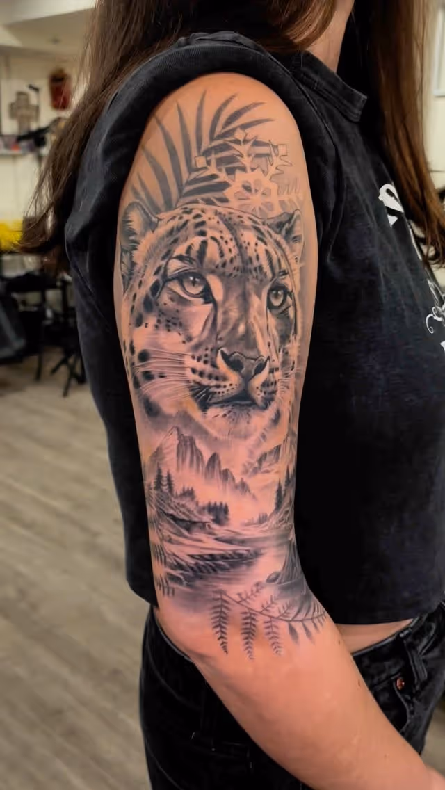 Black and gray leopard tattoo with mountains, trees, and ferns on upper arm.
