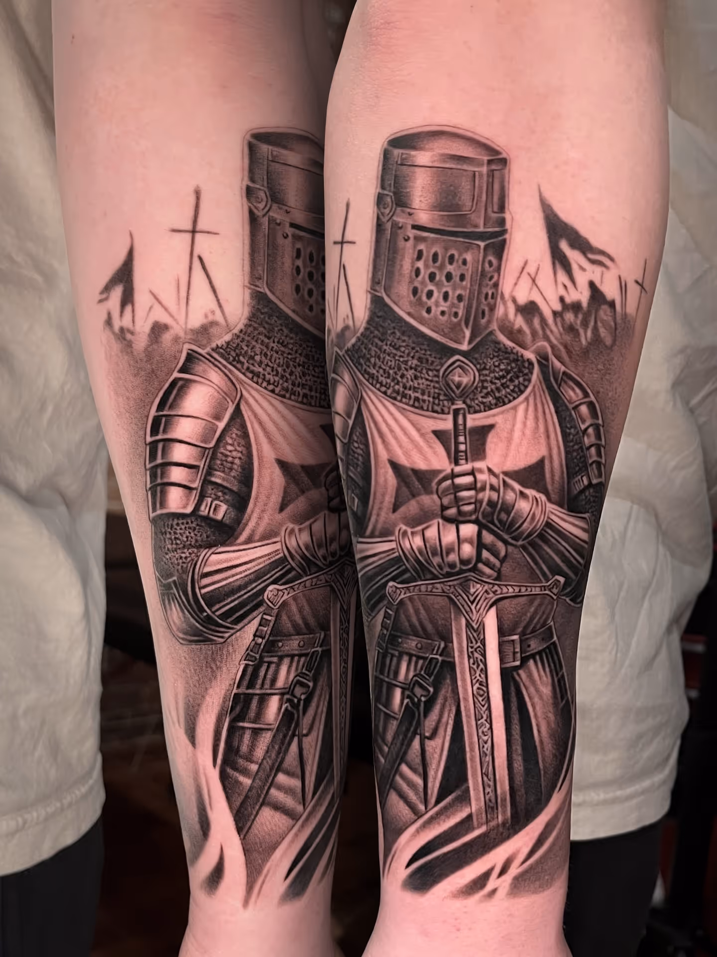 Black and gray forearm tattoos of a medieval knight in armor holding a sword, with crosses in the background