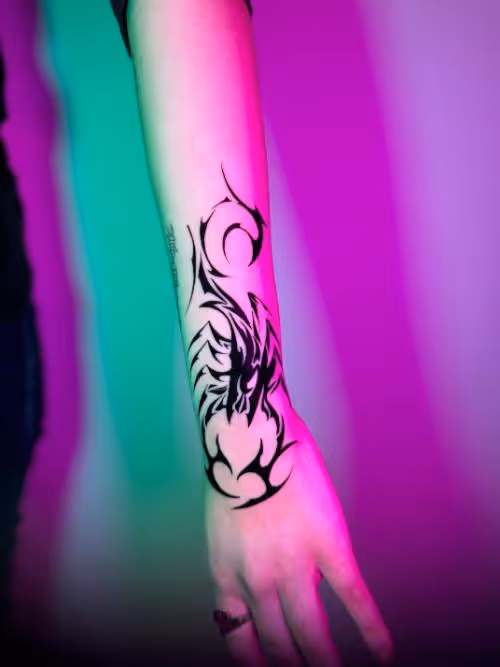 Black tribal dragon tattoo on a forearm under pink and green neon lighting.