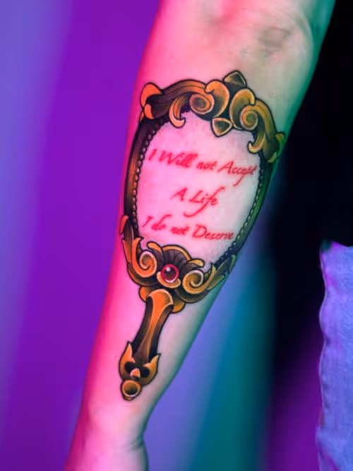 Forearm tattoo of an ornate hand mirror with red script reading I will not accept a life I do not deserve.
