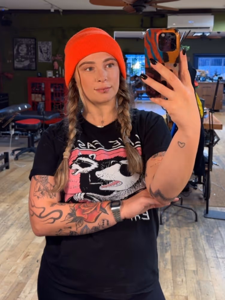 Tattooed person in an orange beanie with braids taking a mirror selfie in a tattoo studio.