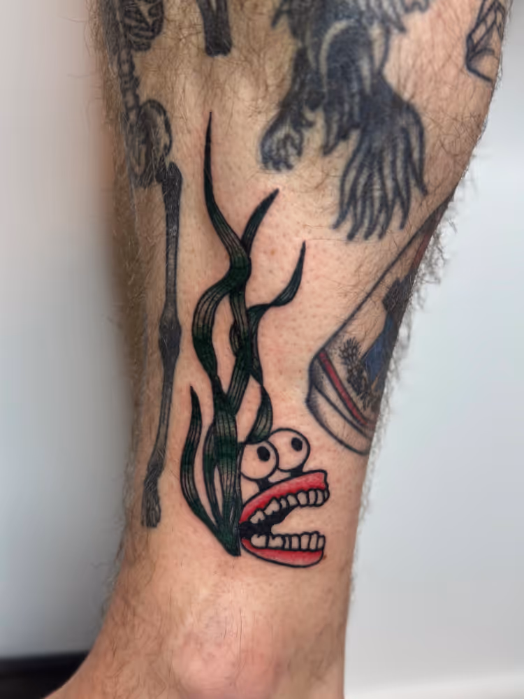 Cartoon seaweed monster with big eyes and teeth tattooed on a calf