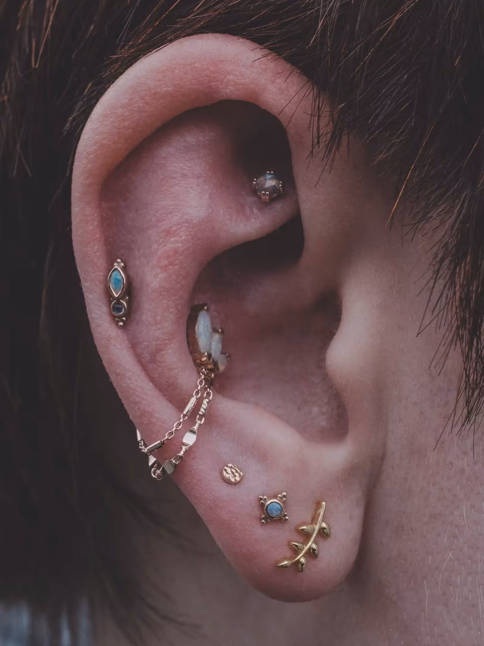Multiple gold and opal ear piercings with chain detail on helix, conch, and lobe.