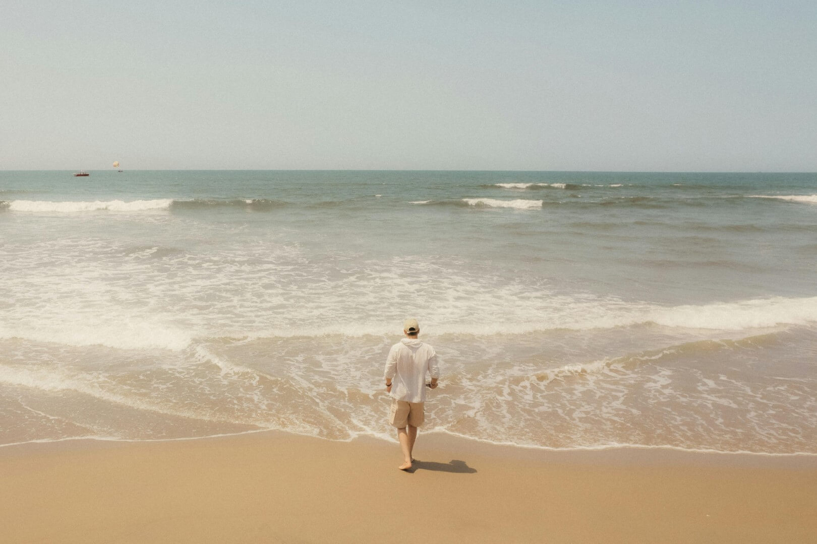 A person in a white shirt and shorts walks barefoot toward the gentle waves of a calm, sandy beach under a clear sky.