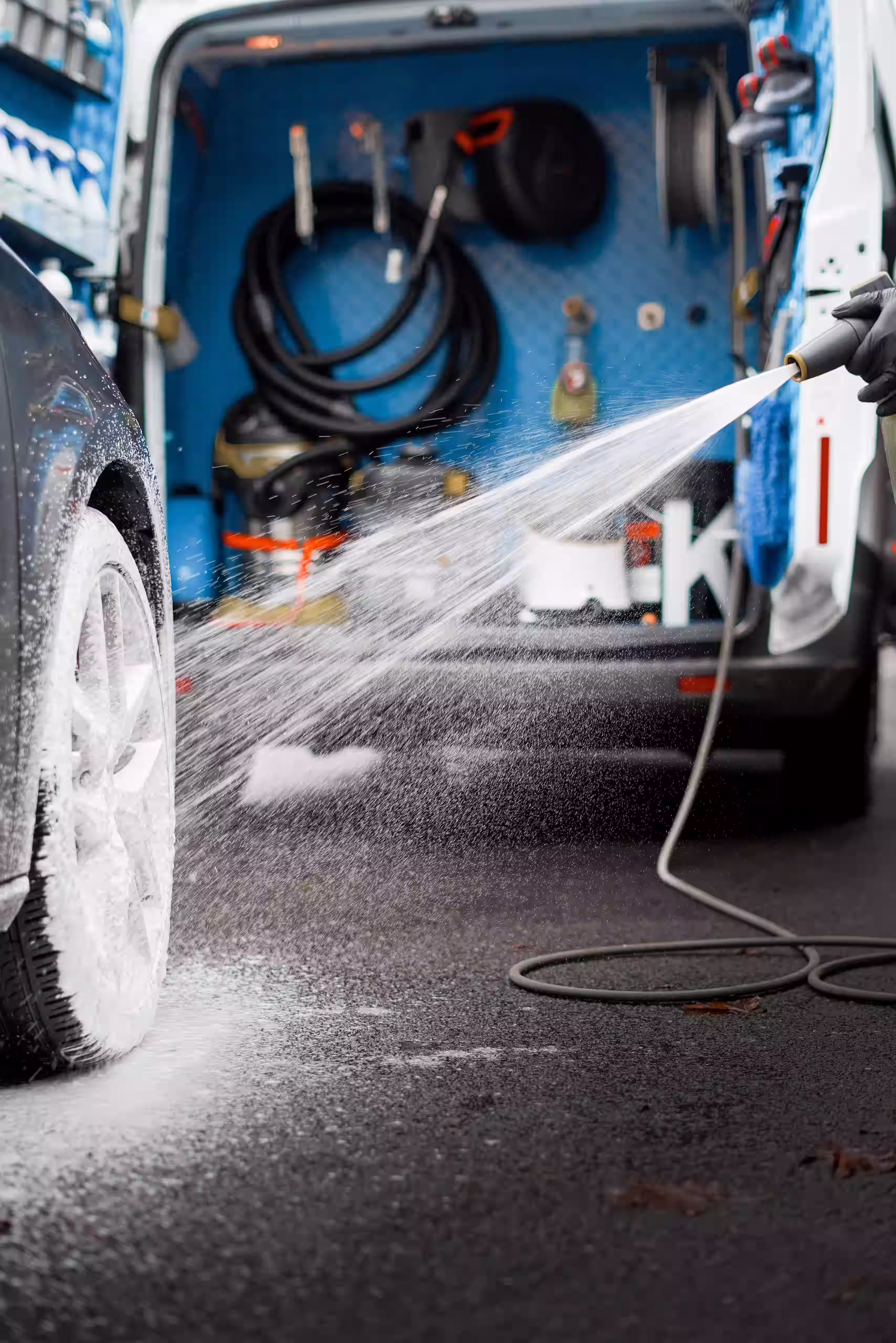 Person using a hose to spray foam on a car tire outside a van equipped with cleaning supplies.