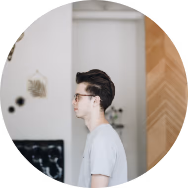 Profil d'un jeune homme portant des lunettes, debout dans une pièce avec un mur clair et des décorations murales.