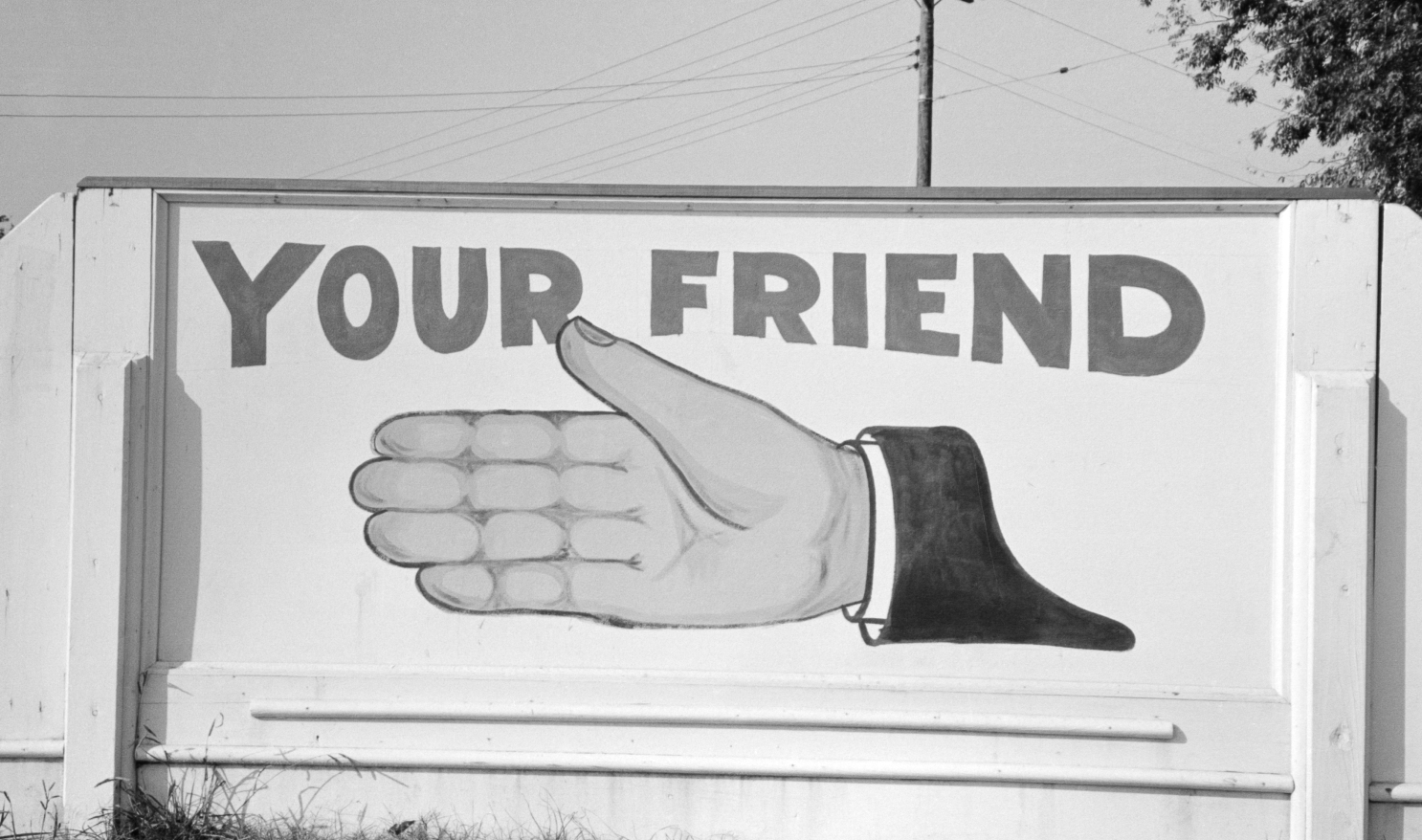 A large black-and-white mural featuring a hand with palm open and the text "YOUR FRIEND" above it, mounted on a sign. The background displays utility poles and trees.