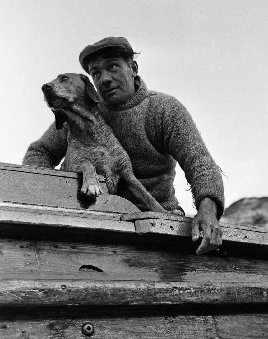 A man in a wool sweater and cap sitting on a wooden boat, looking thoughtfully into the distance while a dog leans against him. The image is in black and white, capturing a serene moment at sea.