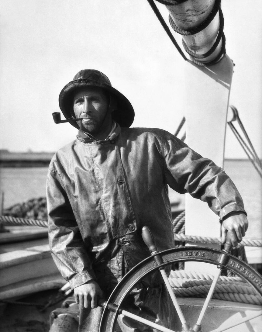 A man in a raincoat and hat stands at the helm of a boat, holding the ship's wheel, with water and ropes visible in the background.
