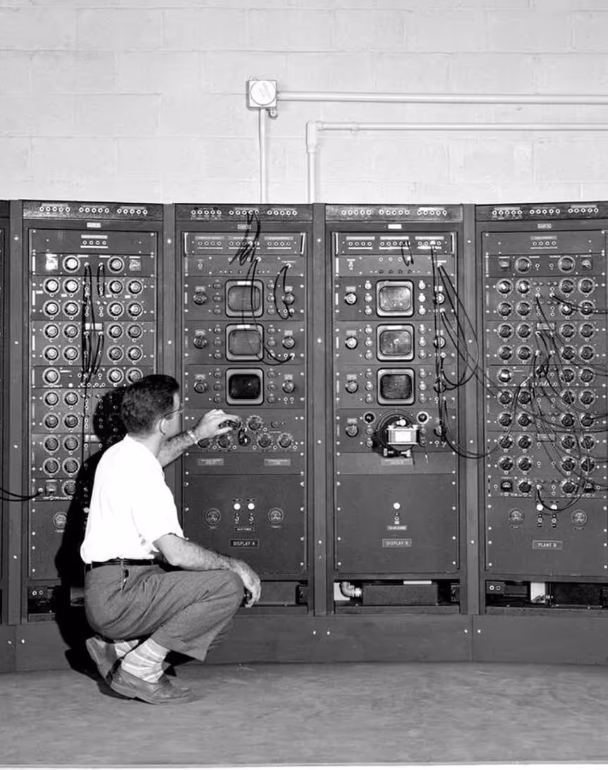 A man adjusting controls on a large bank of vintage electronic equipment in a laboratory setting. The panels have various knobs and dials, and the man is kneeling on the floor while interacting with the machinery.