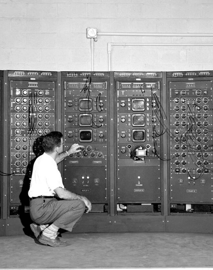 A man adjusting controls on a large bank of vintage electronic equipment in a laboratory setting. The panels have various knobs and dials, and the man is kneeling on the floor while interacting with the machinery.