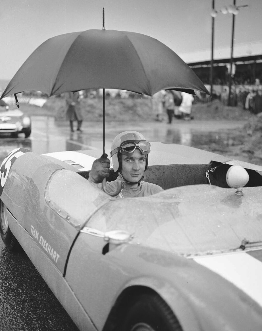 A race car driver sitting in a vintage sports car, holding a red umbrella. The driver wears goggles and a cap, while wet pavement and a racetrack crowd are visible in the background.