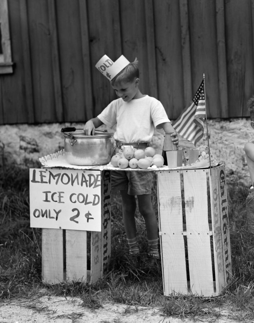 A young boy wearing a paper hat stands at a makeshift lemonade stand, stirring a pot. On the table, there is a sign saying "Lemonade Ice Cold Only 2¢" alongside a small American flag and some lemons.