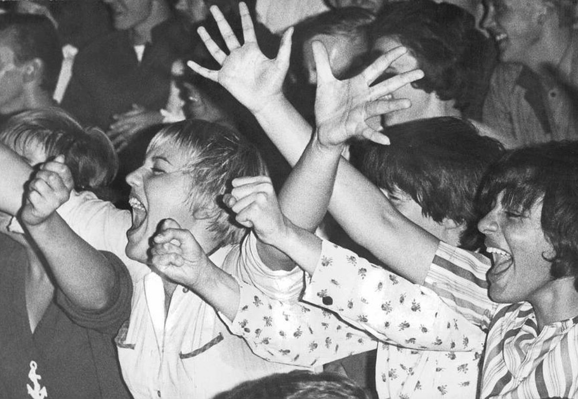 A crowd of enthusiastic fans raising their arms and cheering at a concert or event, captured in black and white. Several individuals are visibly excited, some with wide smiles, showing a strong emotional response.