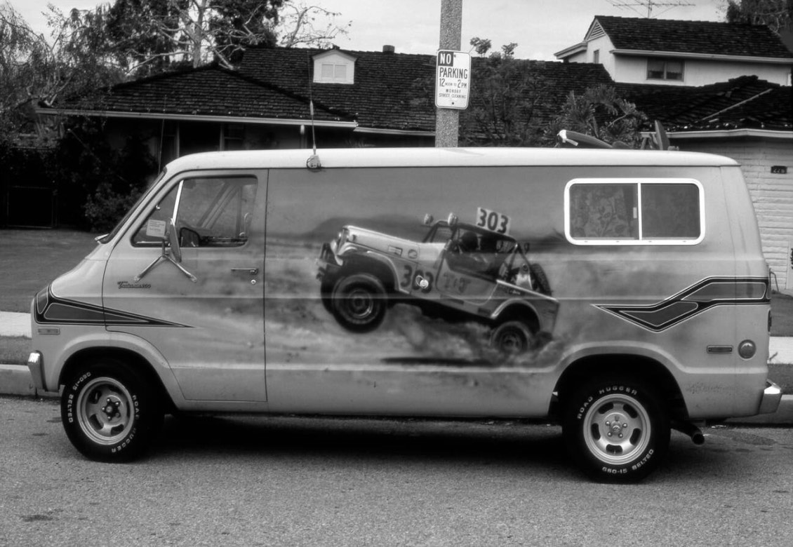 A vintage van with a dynamic mural featuring a racing car, depicting it in mid-air while kicking up dust. The van is parked on a street with a house in the background and a no parking sign visible.
