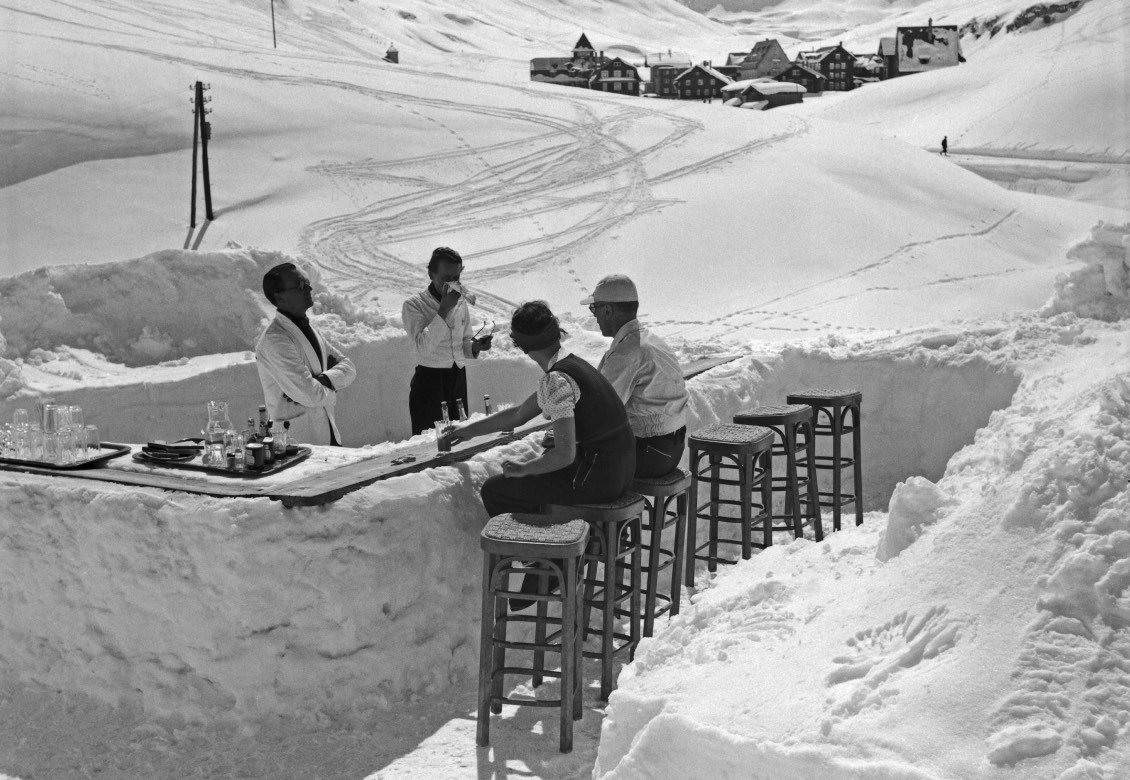 A bar made of snow with four people sitting on stools, enjoying drinks. Snow-covered mountains in the background and a village visible in the distance under a clear sky.