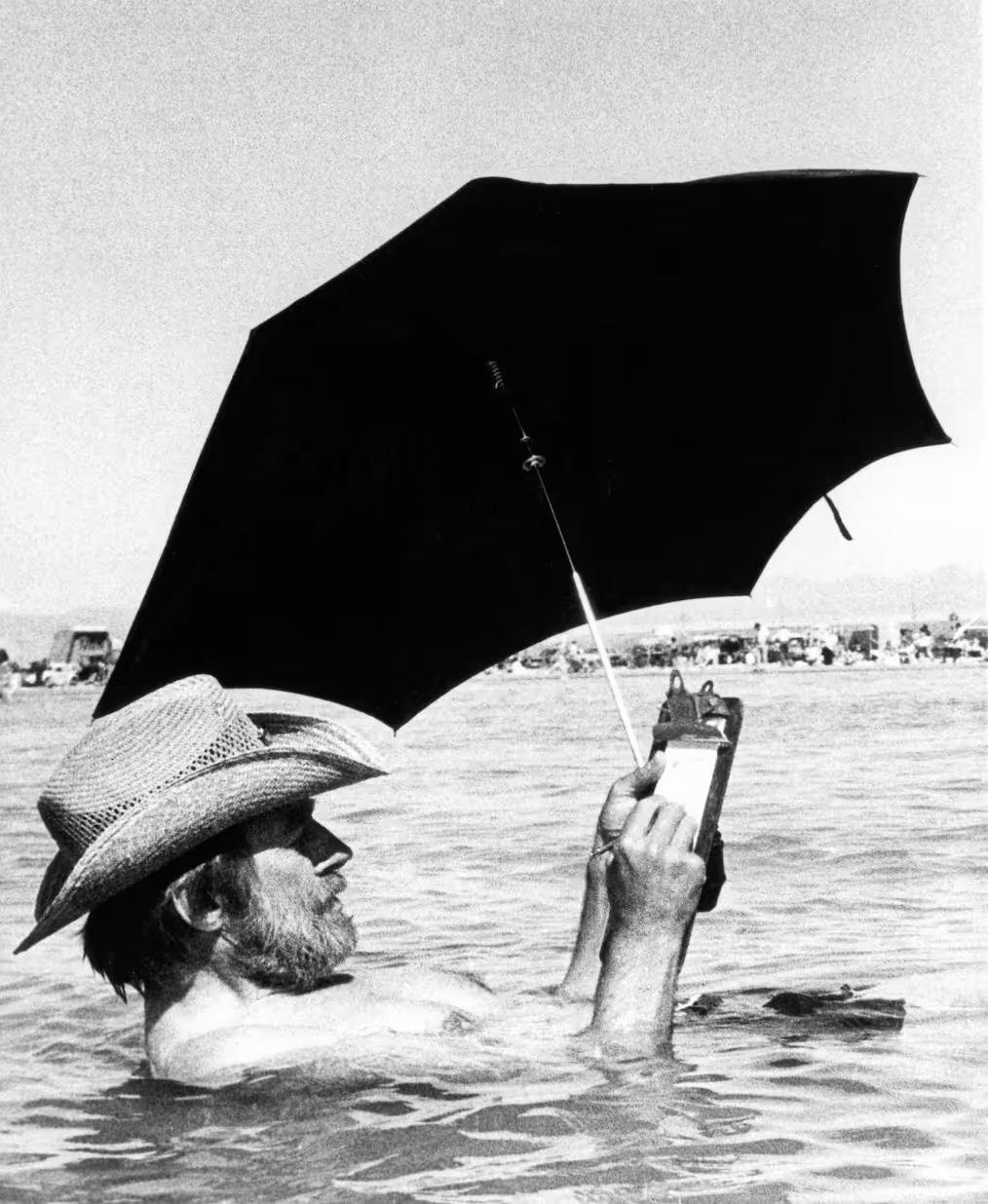 A man relaxing in water while holding a clipboard and a black umbrella for shade. He wears a straw hat and is partially submerged in the water, with a beach scene visible in the background.