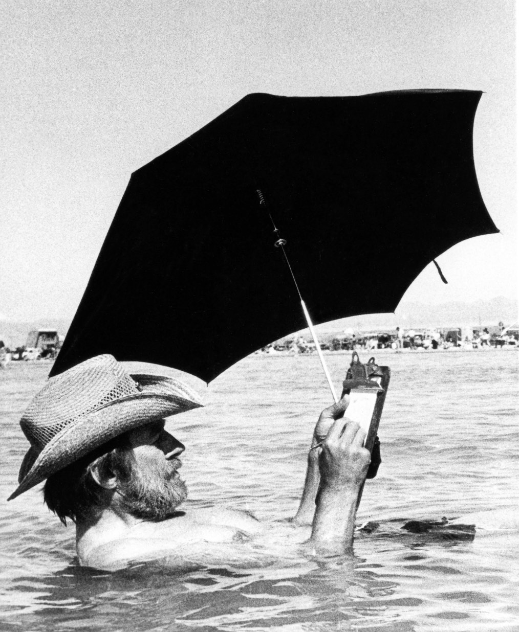A man relaxing in water while holding a clipboard and a black umbrella for shade. He wears a straw hat and is partially submerged in the water, with a beach scene visible in the background.