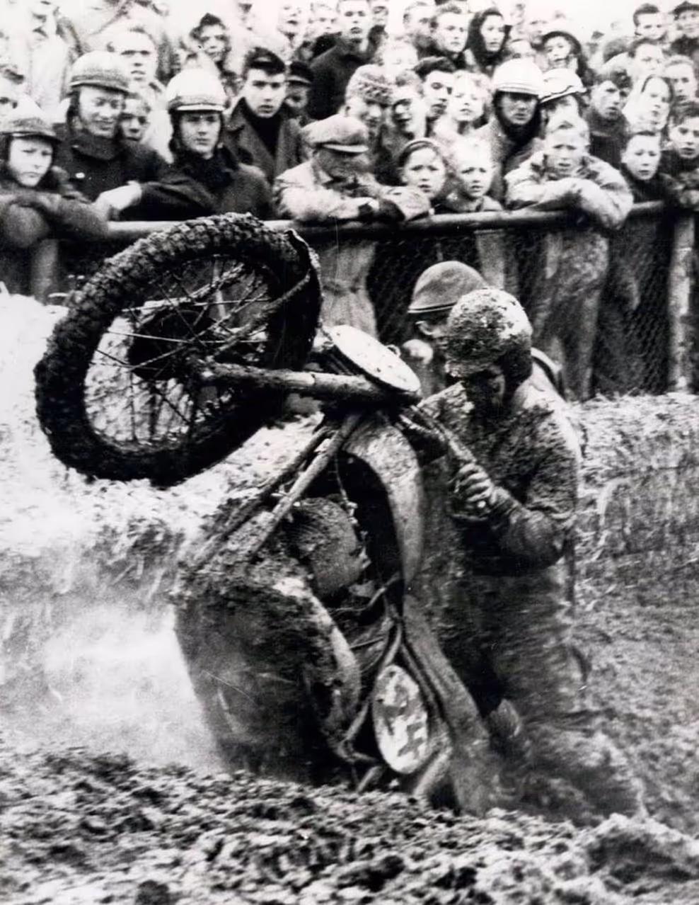 A motorcycle racing event where a rider is struggling to control a fallen bike in a muddy area, with a crowd observing the situation in the background.