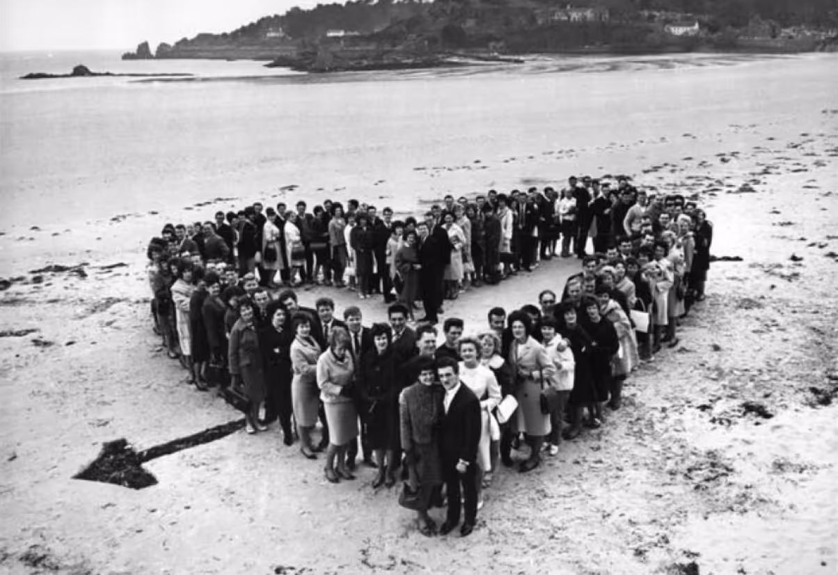 A large group of people arranged in the shape of a heart on a beach, with the ocean visible in the background. The scene is captured in black and white, highlighting the group and the shoreline.