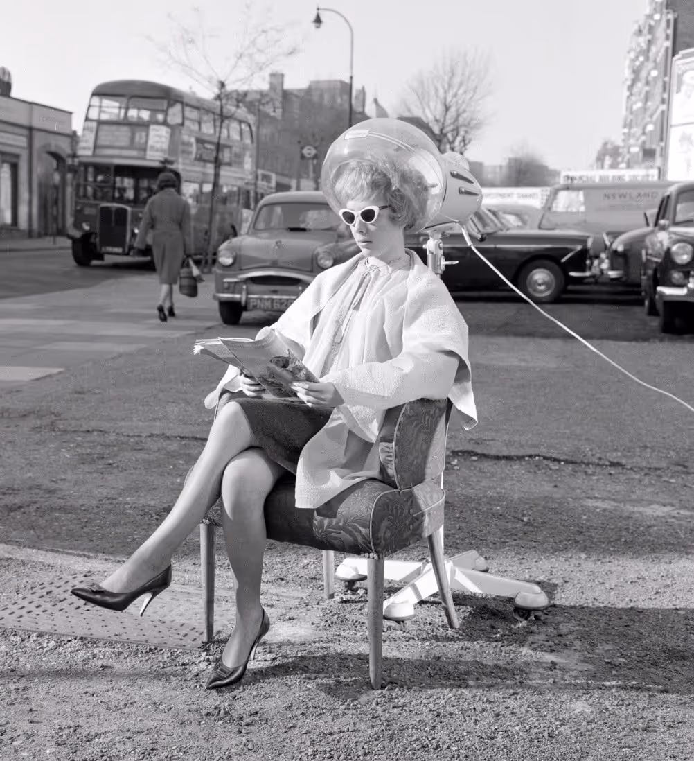 A woman sitting in a chair outside, wearing sunglasses and a hairdryer helmet, reading a magazine. Vintage cars and a double-decker bus are visible in the background, depicting a lively street scene.
