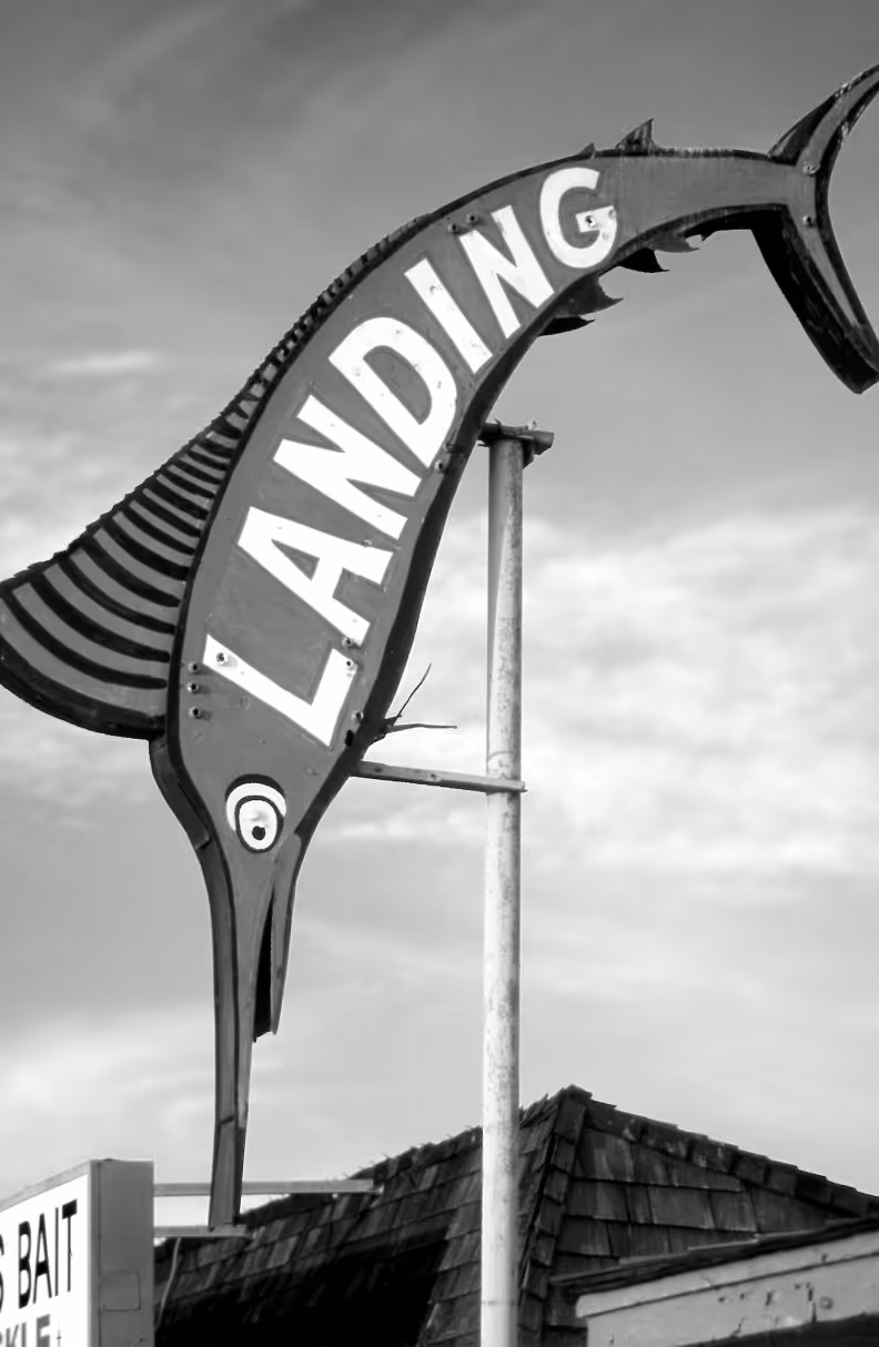 A large sign shaped like a marlin fish with the word "LANDING" prominently displayed. The scene is in black and white, showcasing a cloudy sky in the background and part of a building roof below.