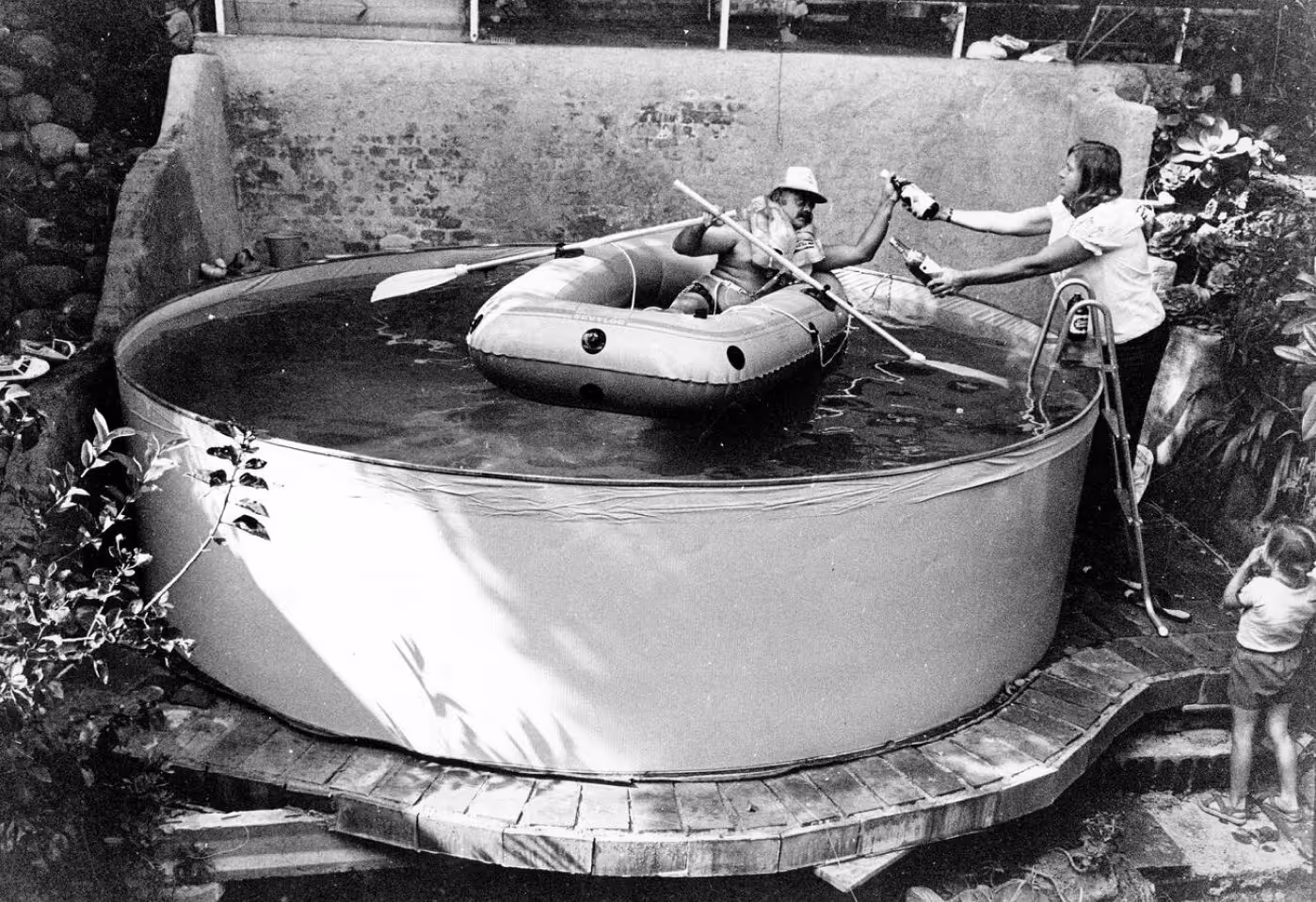 A black and white photo of a circular pool with a person in a small inflatable raft paddling. Another person stands outside the pool, reaching out to hand something to them. A child is visible nearby, observing the scene.