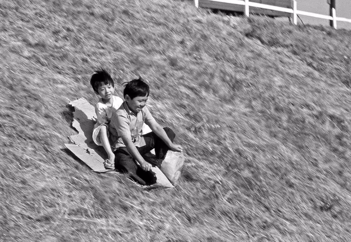 Two children sitting on a makeshift sled made of cardboard, sliding down a grassy slope. One child is in front with a focused expression, while the other child is seated behind, looking back towards the camera.