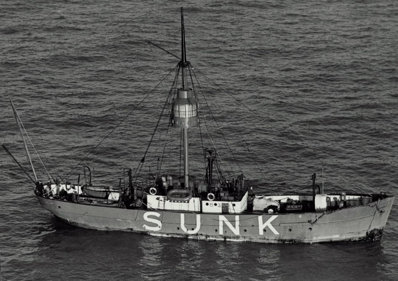 An old ship with the word "SUNK" painted on its side, partially submerged in water. The ship features a tall mast and various equipment on deck, surrounded by calm ocean water.