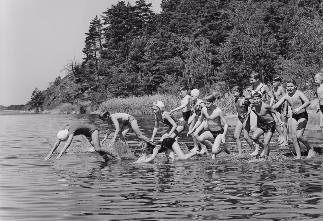 A group of children in swimsuits, some diving and others standing at the edge of a river, surrounded by trees. The scene captures a lively summer day outdoors.