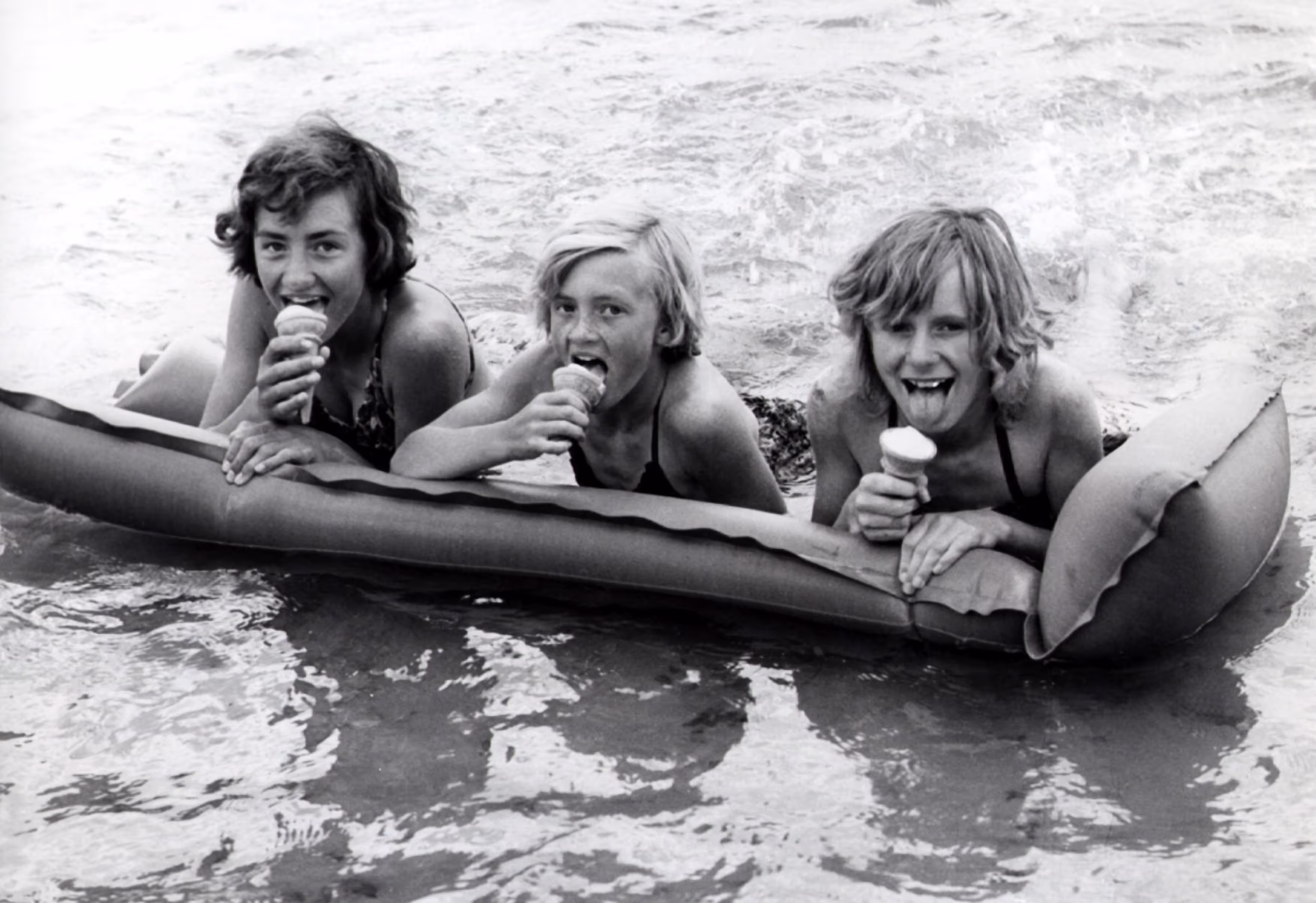 Three children on a floating raft in the water, each holding an ice cream cone. They are wearing swimsuits, and the scene is in black and white.