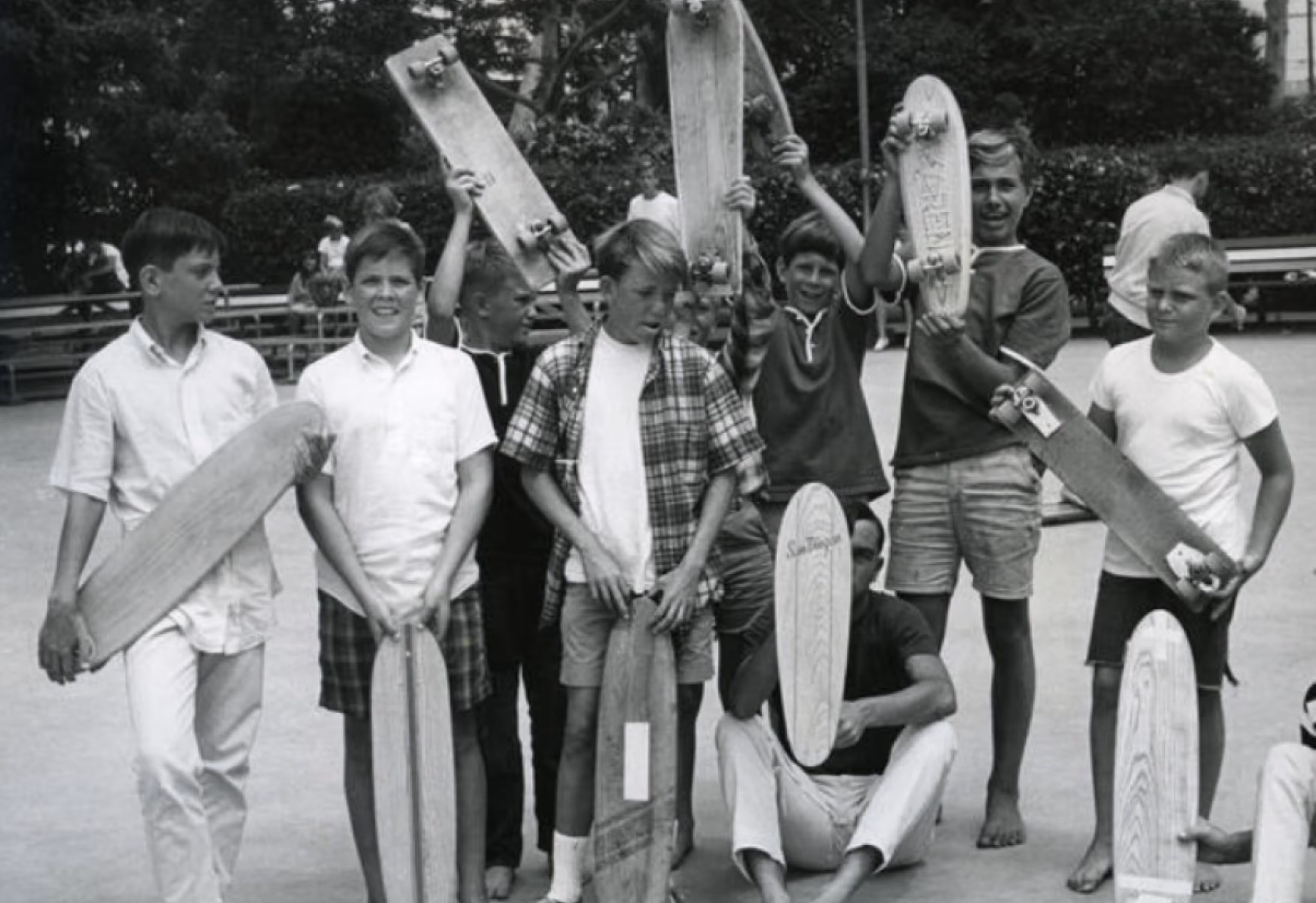 A group of children and teenagers gathered outdoors, holding various skateboards. Some are posing with their skateboards above their heads while others stand casually. The background features benches and greenery.