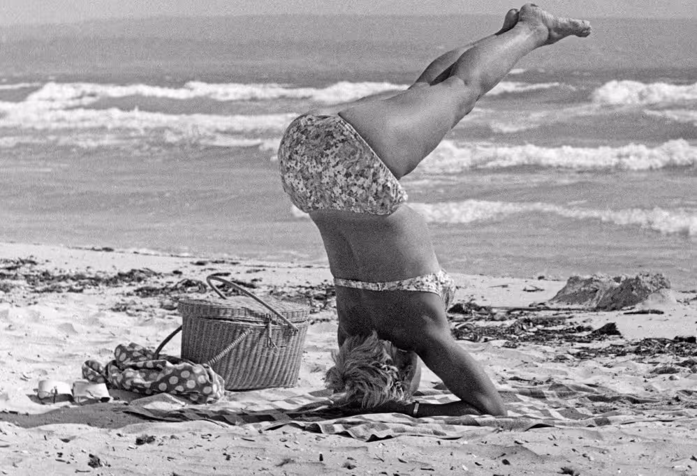 A person in a swimsuit is performing a headstand on a beach towel while on a sandy beach. A picnic basket and other items are nearby, with ocean waves visible in the background.