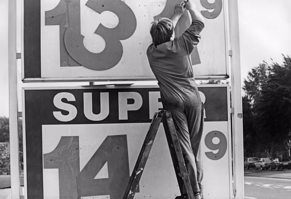 A person adjusting numbers on a large outdoor sign while standing on a ladder. The sign displays the numbers '13' and '14' along with the word 'SUPER' in bold letters. The image is in black and white.