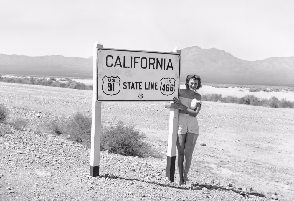 A woman in shorts stands next to a sign indicating the California state line, with desert terrain and mountains visible in the background.