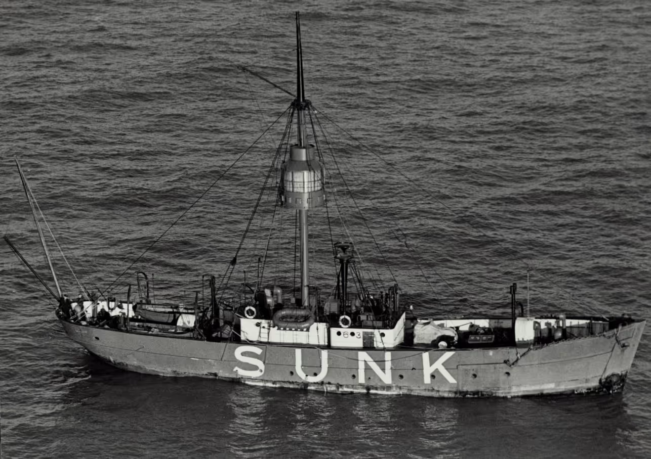 An old ship with the word "SUNK" painted on its side, partially submerged in water. The ship features a tall mast and various equipment on deck, surrounded by calm ocean water.