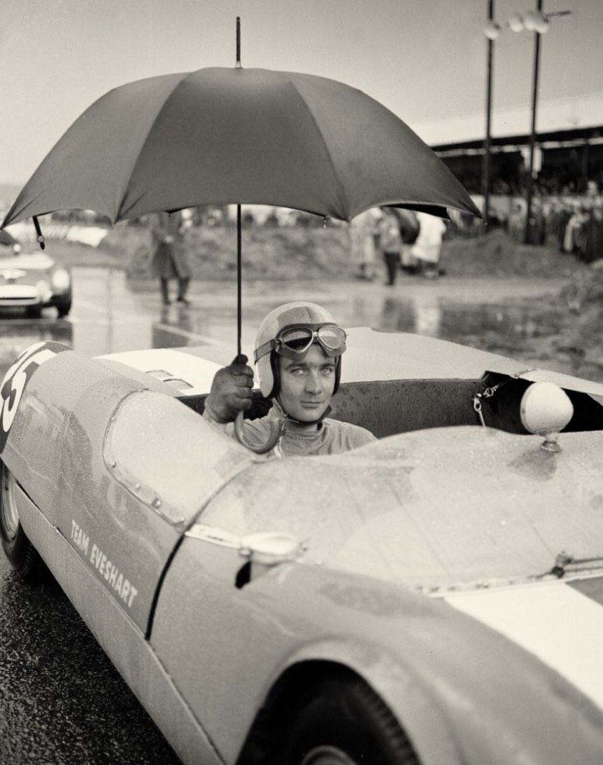 A race car driver sitting in a vintage car holding an umbrella while waiting on a rainy racetrack. The driver is wearing goggles and a helmet, with spectators and other vehicles in the background.