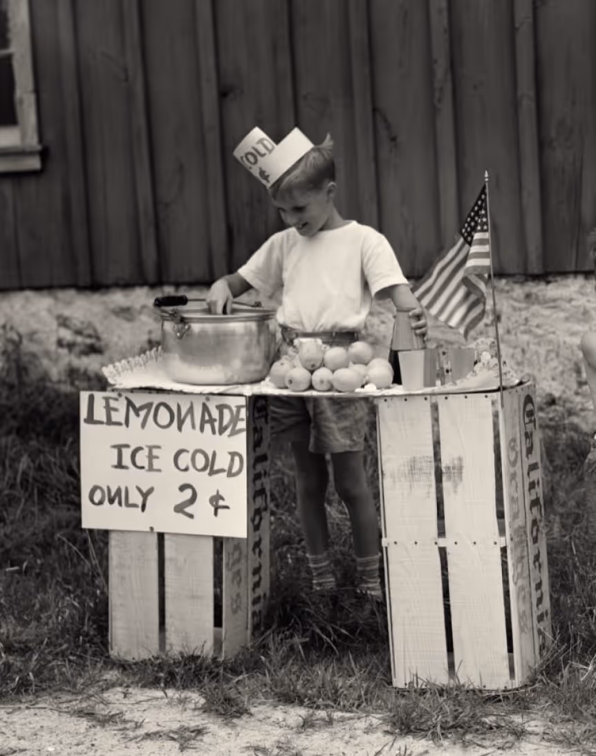 A young boy wearing a paper hat stands behind a lemonade stand made from crates, with a sign reading "LEMONADE ICE COLD ONLY 2¢". He is preparing lemonade, with a small American flag on the stand.