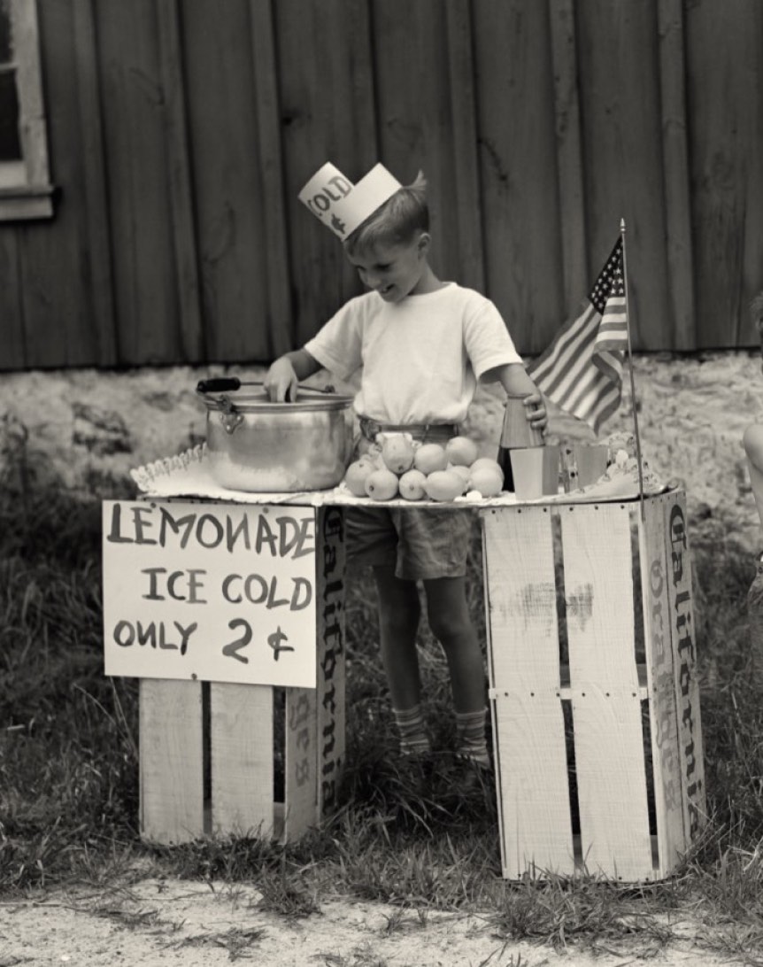 A young boy wearing a paper hat stands behind a lemonade stand made from crates, with a sign reading "LEMONADE ICE COLD ONLY 2¢". He is preparing lemonade, with a small American flag on the stand.