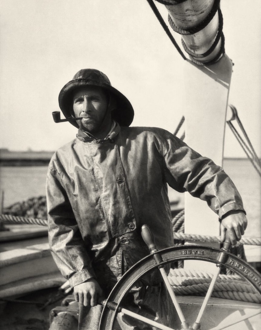 A man wearing a raincoat and a hat stands beside a ship's steering wheel on a boat. The setting includes ropes and the visible water in the background, suggesting a maritime environment.