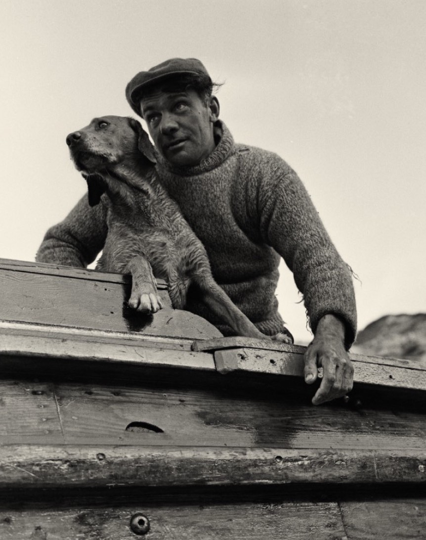 A man wearing a cap and a knitted sweater sitting on the edge of a wooden boat, gazing into the distance, with a dog resting beside him.