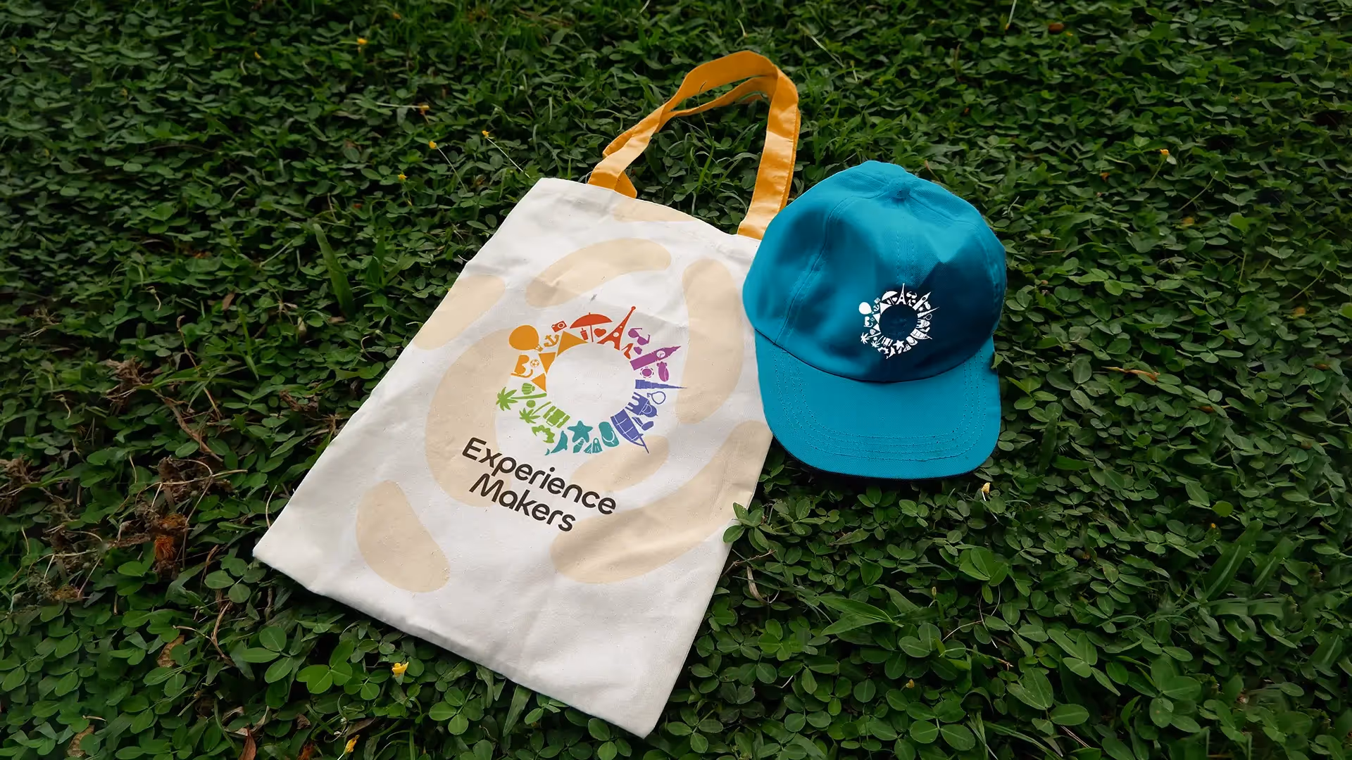 Mockup of tote bag and a baseball cap on some grass, showing the logo of Experience Makers Tourism