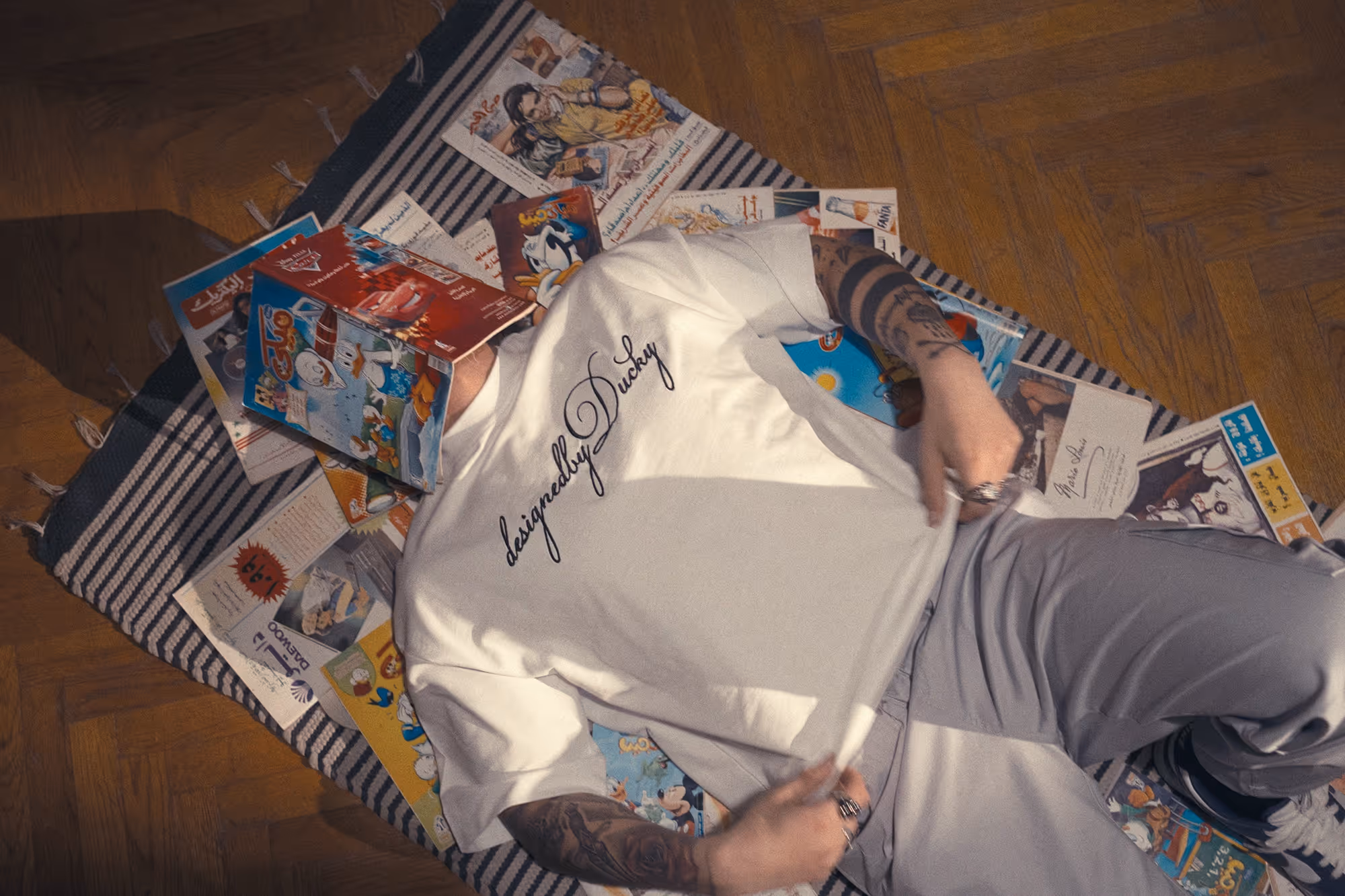 Photo of a guy wearing a Designed by Ducky shirt laying on a carpet with comic books around him and on his face