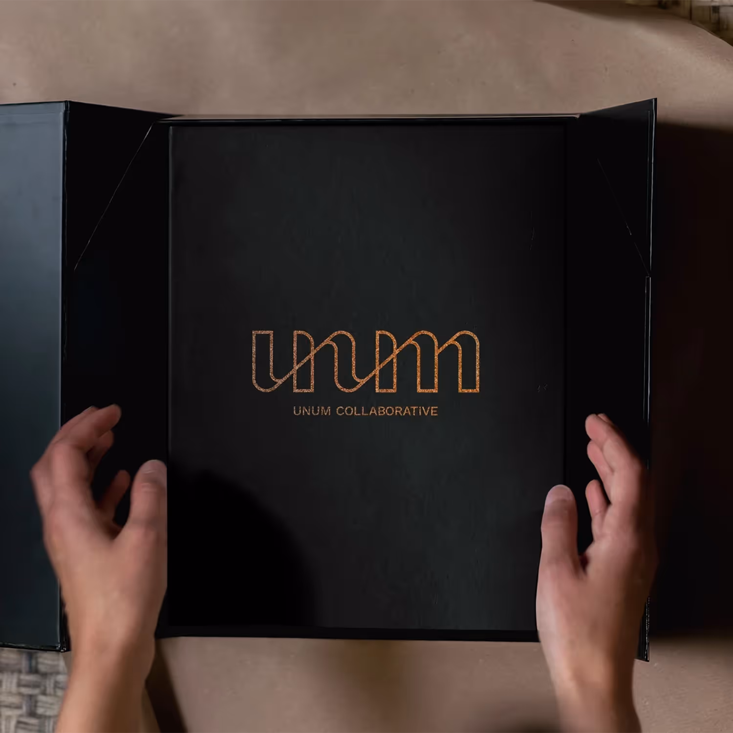 Hands opening a sleek black presentation box with the logo “UNUM COLLABORATIVE” in orange on the centre lid, showcasing elegant packaging design created by a branding agency.