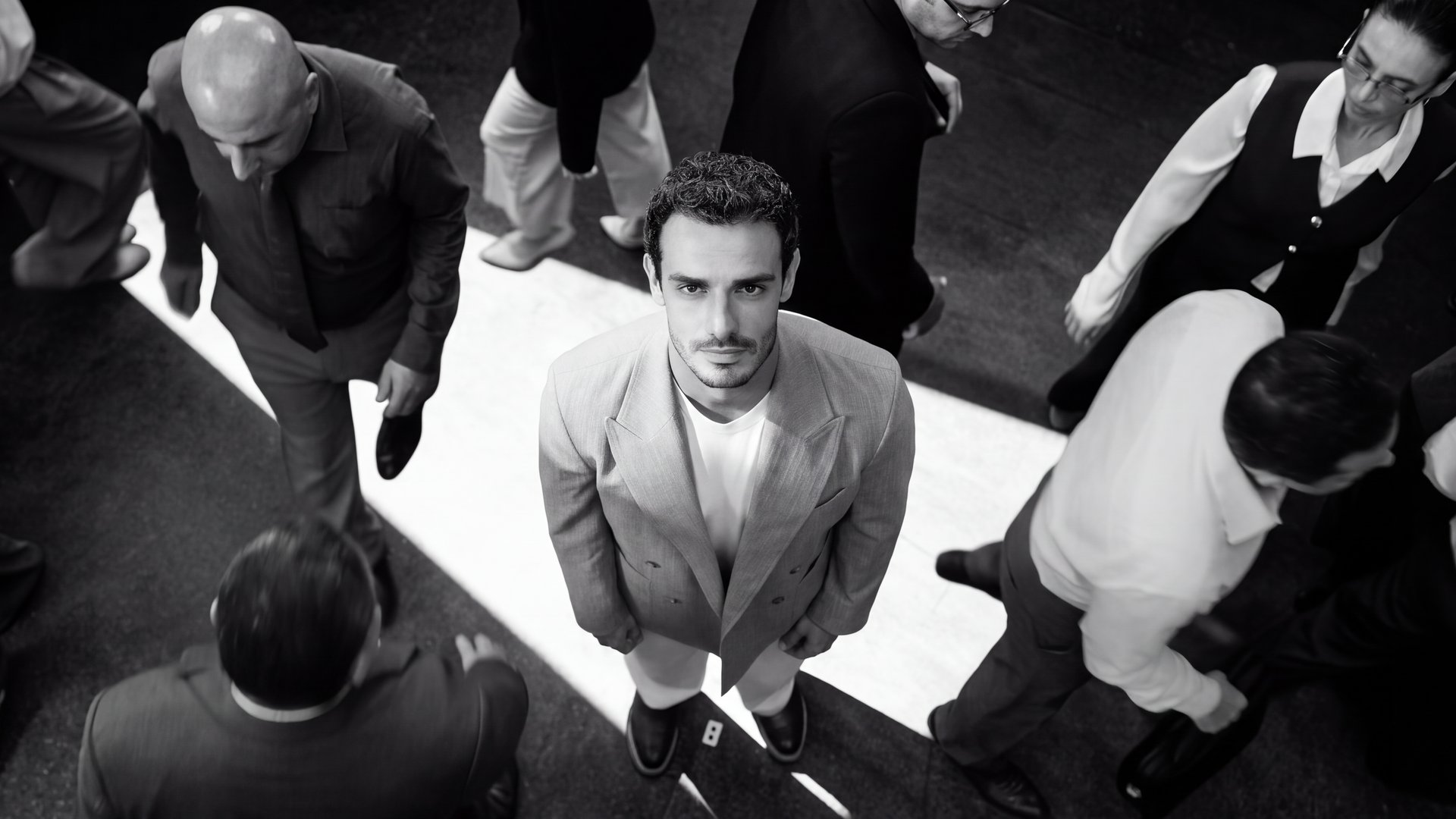 Overhead shot of the lead standing still in a pool of light as a crowd moves around him