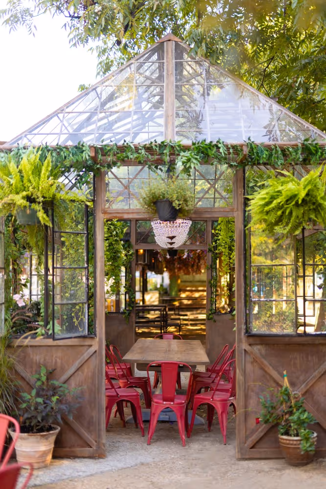 Glass greenhouse with red chairs, hanging plants, and wooden dining table