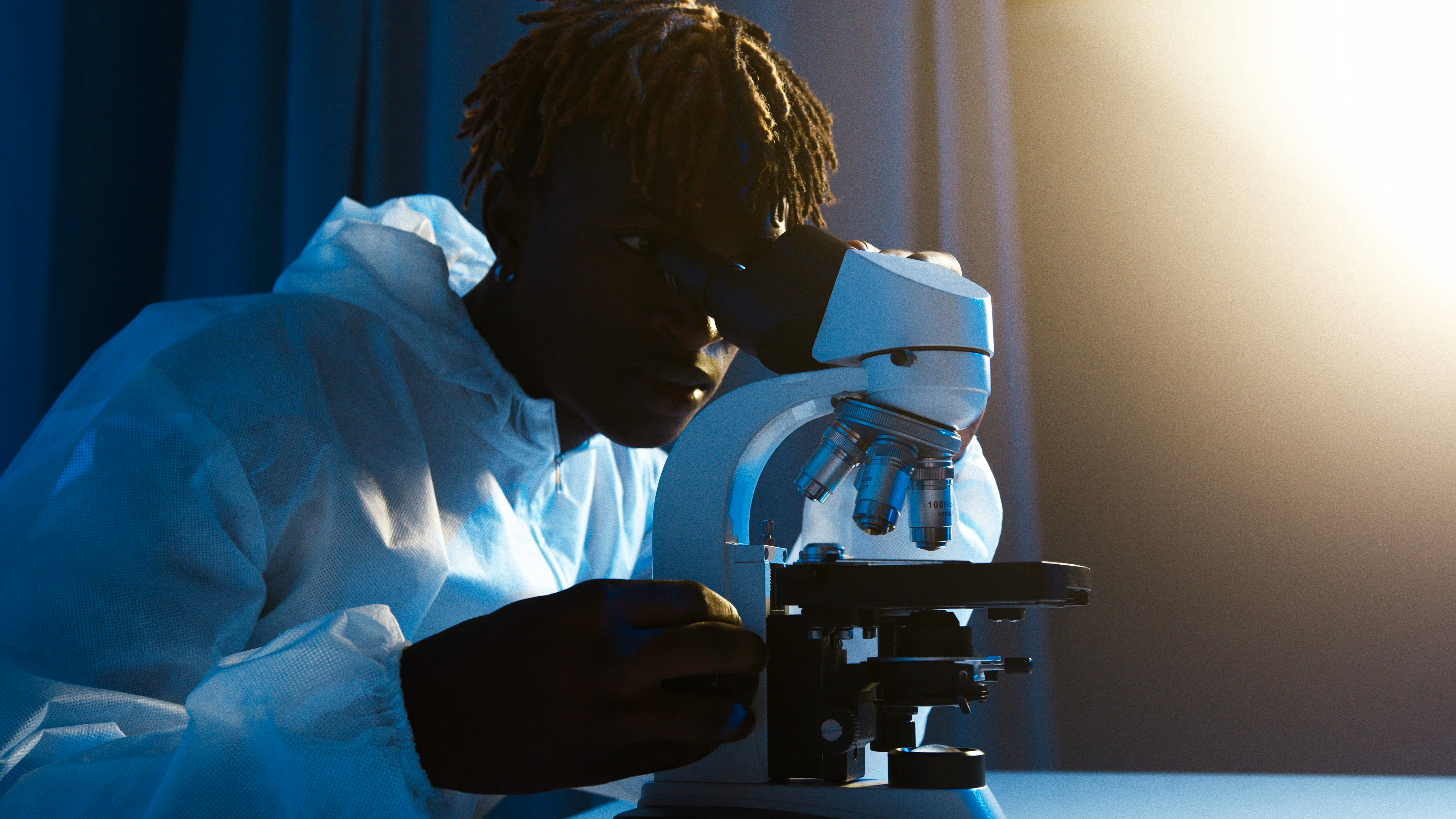 Scientist in protective suit looking into a microscope under focused lighting.