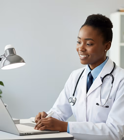 Smiling female doctor wearing a white coat and stethoscope, working on a laptop at a desk with a lamp.