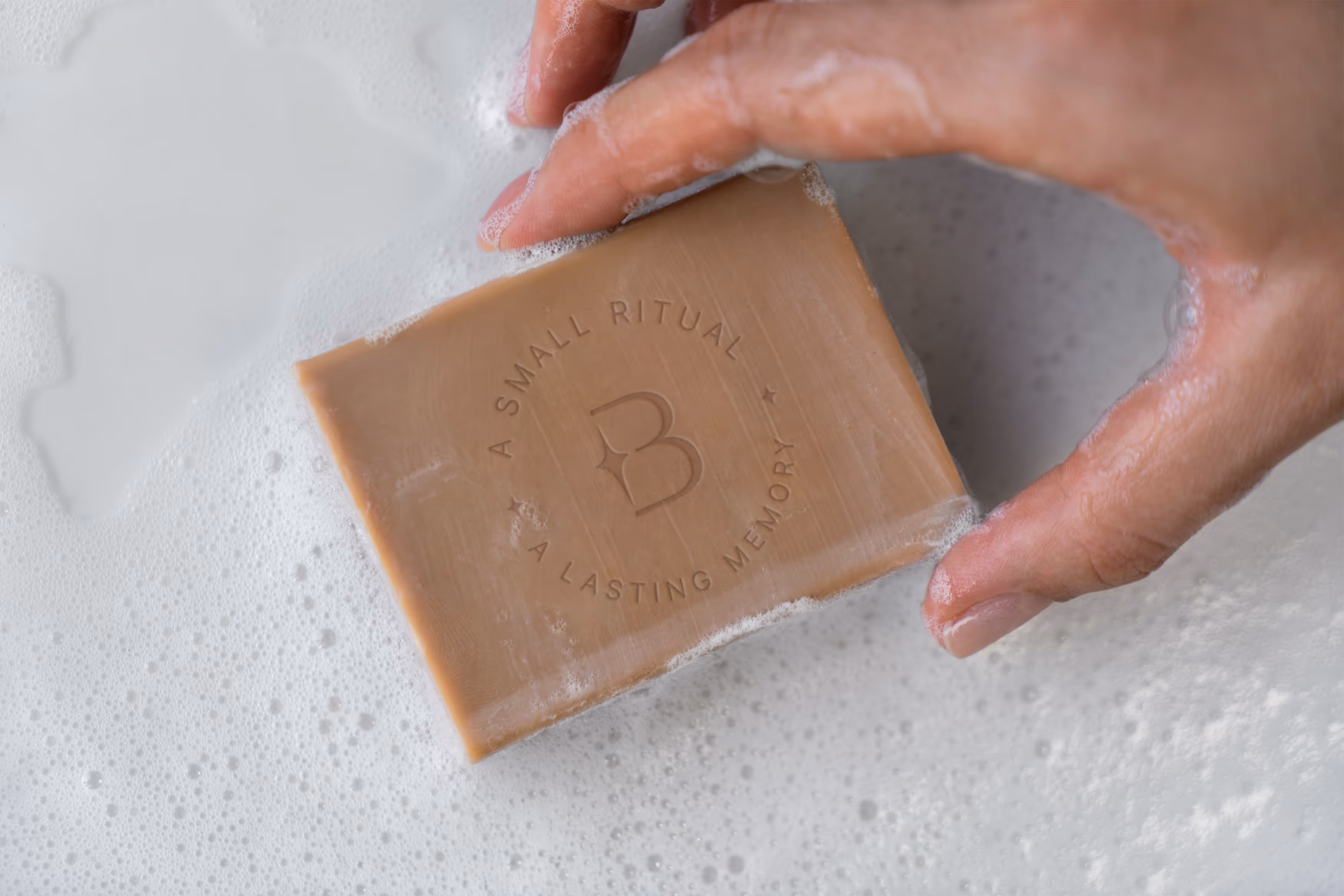 A hand holding a bar of soap above foamy water, embossed with the Star Baths logo and the phrase 'A Small Ritual • A Lasting Memory'.